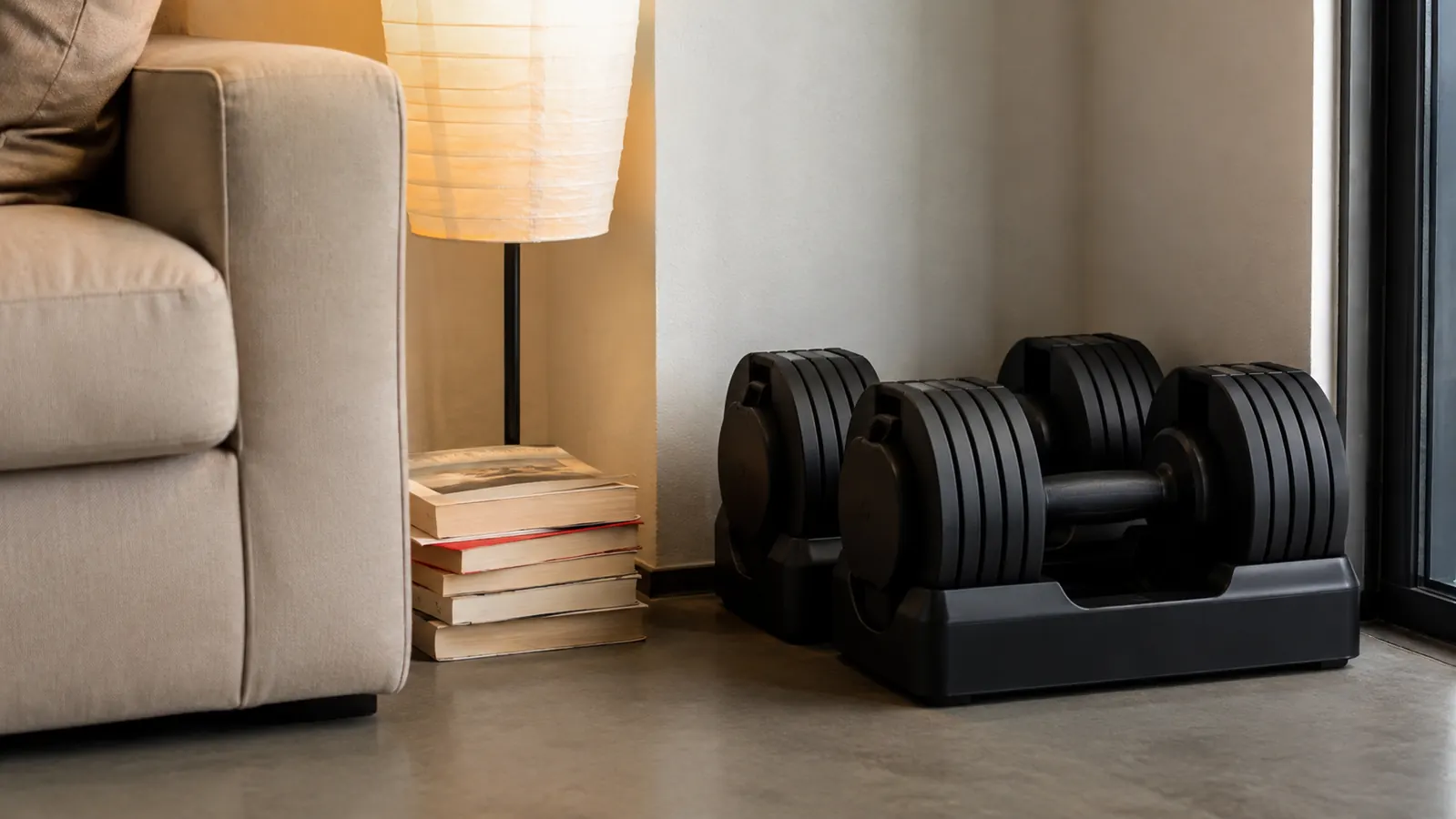 Adjustable dumbbells stored neatly in a corner next to a floor lamp and a stack of paperback books, showing how little space they take up