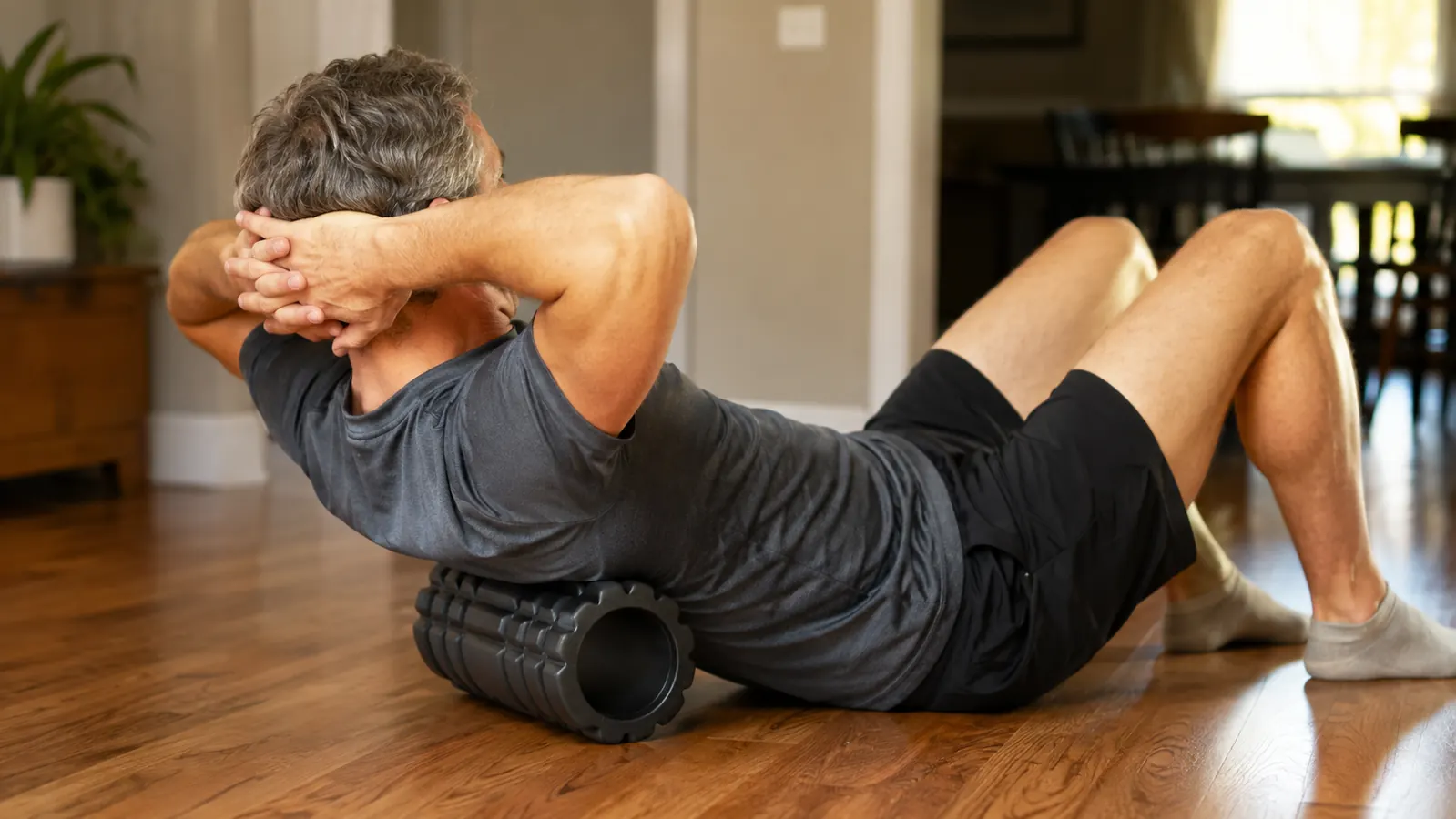 A person sitting on a hardwood floor using a foam roller on their upper back, leaning back against it with hands behind head, natural light